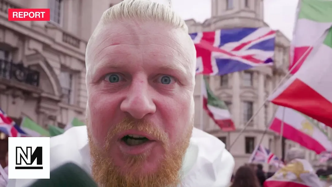 A still from a Novara report: a man speaking to camera in a street full of flags.